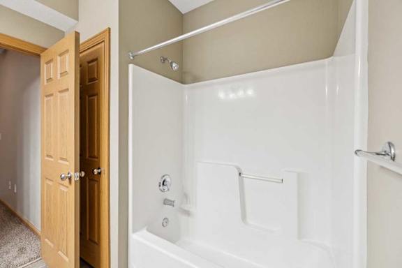 Tub and shower combination with built-in surround, towel bar, and wood-trimmed door leading to the hallway.
