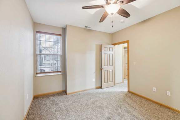 Secondary bedroom with plush carpet, ceiling fan, window with blinds, and wood-trimmed door.