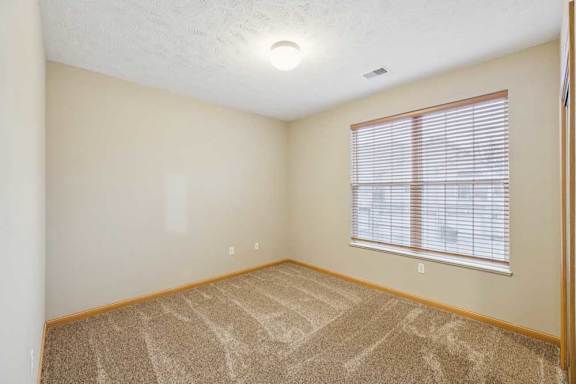Bright bedroom with large window and blinds, neutral carpet, and wood trim for a clean, updated apartment interior.