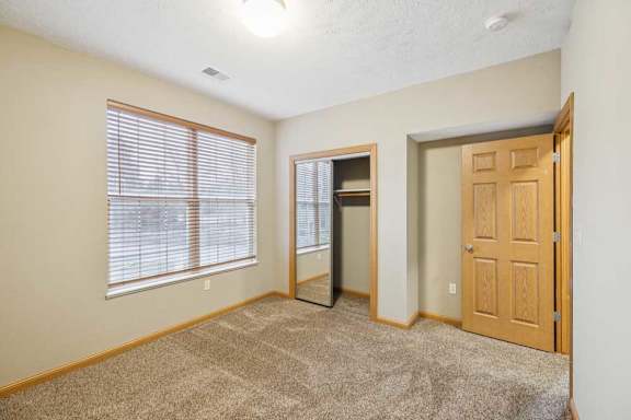 Bedroom featuring a window with blinds, open closet nook, and neutral carpet in a well-lit apartment space.