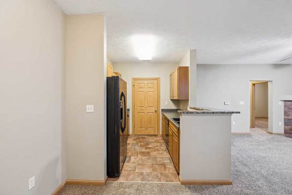 Galley-style apartment kitchen with wood cabinets, black refrigerator, laminate countertops, and tile flooring, opening to a carpeted living area.