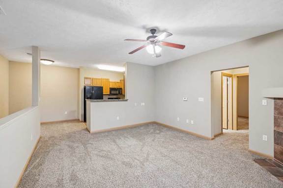 Bright living area with plush carpet, ceiling fan, fireplace with mantel, and large window with blinds.