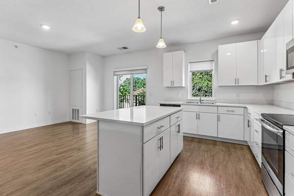 Sleek white kitchen centered around a spacious island with pendant lighting, abundant cabinetry, and windows framing leafy views.