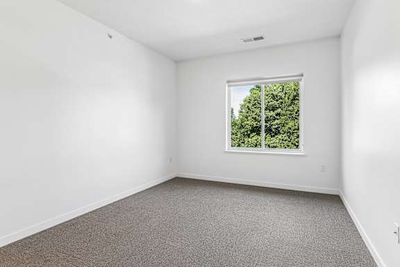 Unfurnished bedroom with soft gray carpeting and a wide window that fills the space with natural light and leafy treetop views.