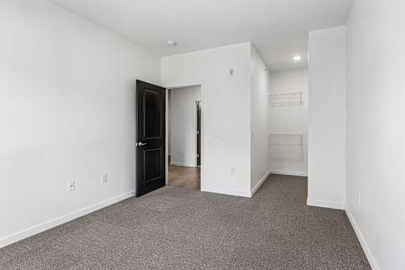 Bright, empty apartment bedroom with gray carpet, white walls, a black door, and an open closet with wire shelving.