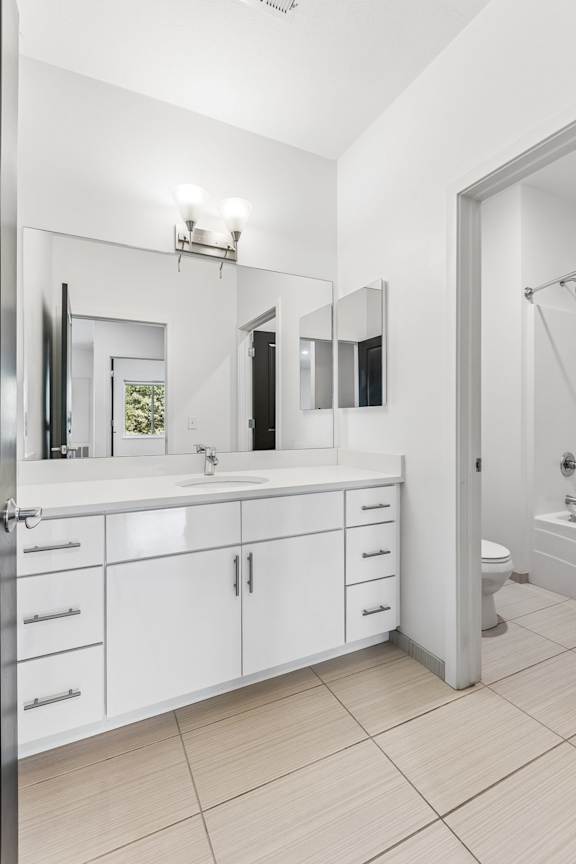 Bright apartment bathroom with a wide vanity, white cabinetry, large mirror, tile flooring, and an adjoining tub and toilet area.