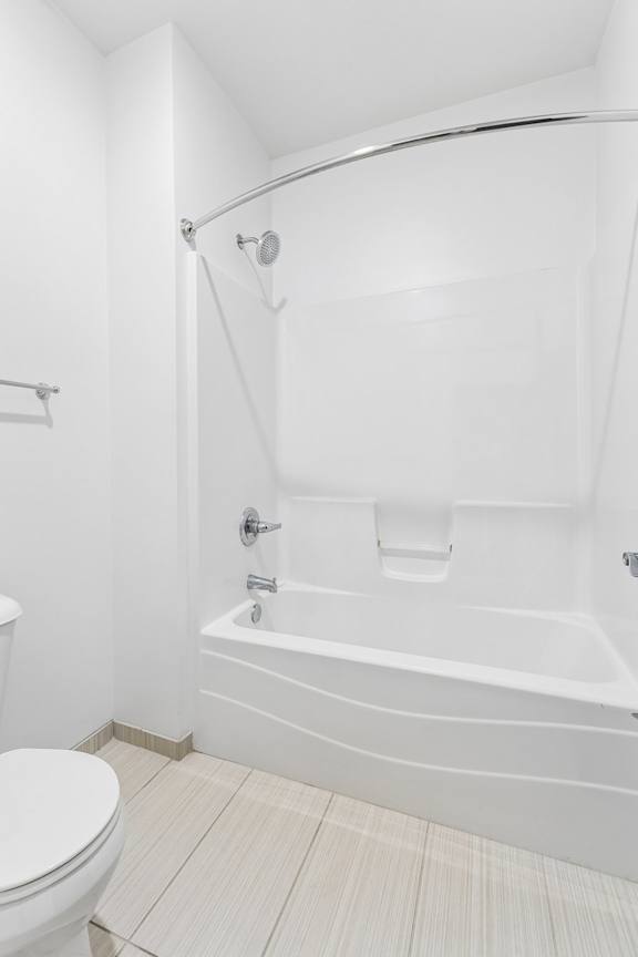Apartment bathroom with a white tub and shower combination, curved curtain rod, built-in shelf, and light tile flooring.