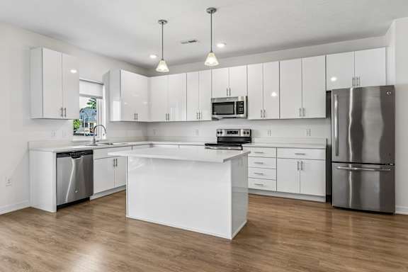 Bright apartment kitchen with white cabinetry, a large center island, stainless steel appliances, pendant lights, and wood-look flooring.