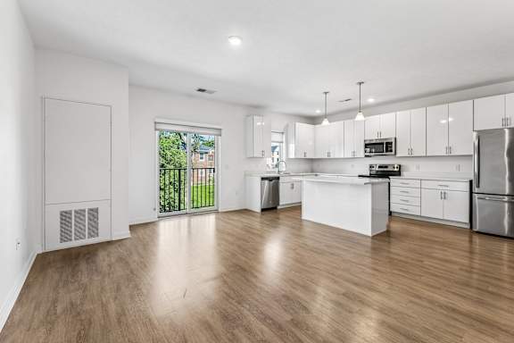 Bright, open apartment kitchen and living area with white cabinetry, center island, stainless steel appliances, and a balcony door.