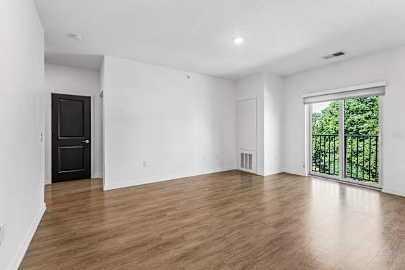 Open living room with warm wood-look flooring, a black interior door, and sliding glass doors opening to leafy outdoor views.