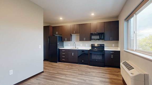 A kitchen with dark wood cabinets next to a living area with hardwood floors and a large window