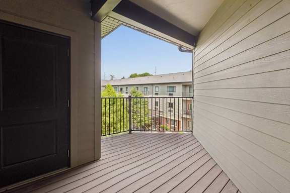 Covered private balcony with wood-look decking and black railings, overlooking the landscaped apartment courtyard and neighboring residences.