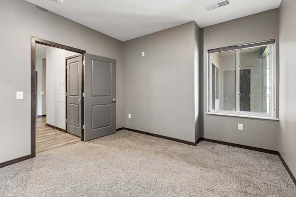 Neutral-toned bedroom with plush carpeting, dark trim, and a large window overlooking the apartment community exterior.