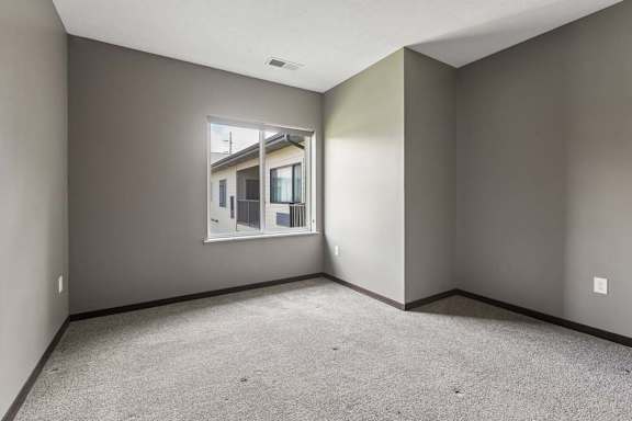 Carpeted bedroom with warm gray walls, dark trim, and a window overlooking a neighboring building within the apartment community.