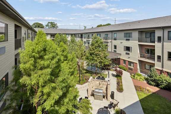 Landscaped apartment courtyard framed by mature trees and residential buildings, with outdoor seating, a grill station, and walking paths.