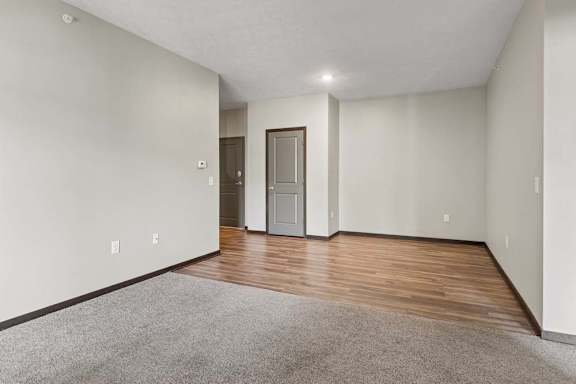 Open living and dining area transitions from soft gray carpet to wood-look flooring, with neutral walls and dark trim.