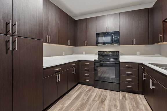Rich espresso cabinetry wraps this apartment kitchen, paired with white counters, black appliances, and wood-look flooring.