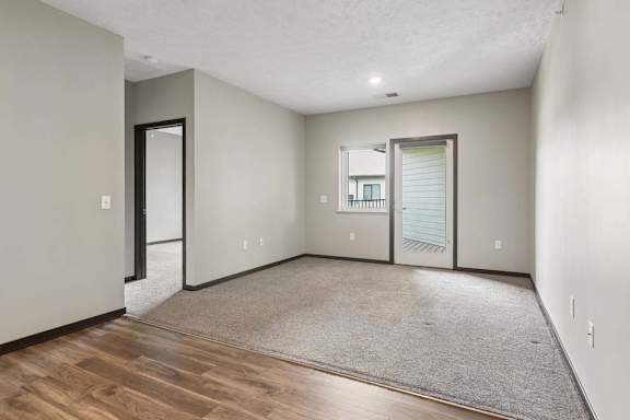 Carpeted living room opens to a private balcony, with neutral walls, dark trim, and wood-look flooring at the entry.