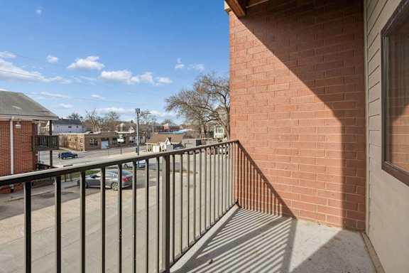 Private apartment balcony with a black metal railing, brick exterior, and neighborhood views under a clear blue sky.