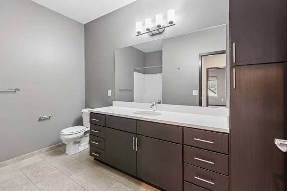 Modern apartment bathroom with a double-sink vanity, dark wood cabinetry, a large mirror, and bright vanity lighting.