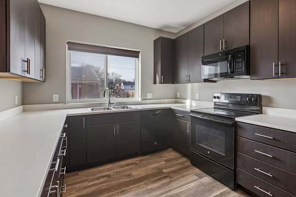 Modern apartment kitchen with dark wood cabinetry, black appliances, white countertops, and a window over the sink.