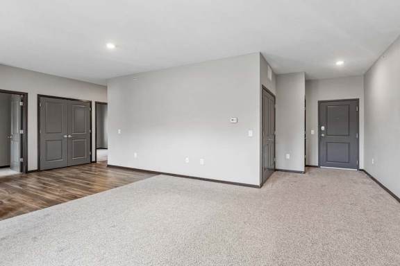 Spacious apartment living room with plush carpeting, recessed lighting, and an open layout connecting to the kitchen and entryway.
