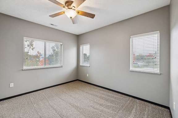 A carpeted bedroom with several windows, and a ceiling fan.