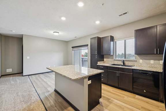A kitchen with dark cabinets, granite counters, a large island, open to a dining room with a sliding glass door to a balcony.