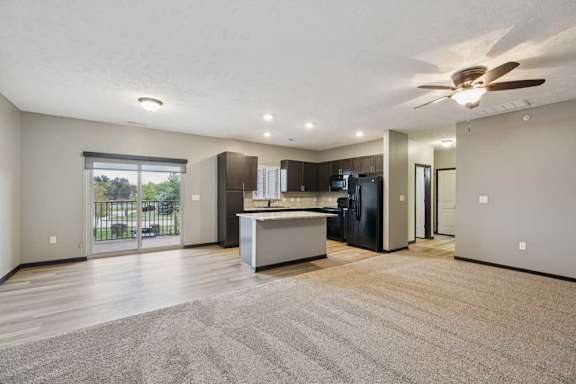 A spacious, carpeted living room open to a modern kitchen with hardwood flooring and dark wooden cabients.