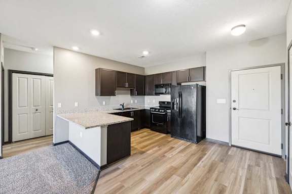 Apartment kitchen with L-shaped counter, dark wood cabinets, black refrigerator and oven, and adjacent entry and storage doors.