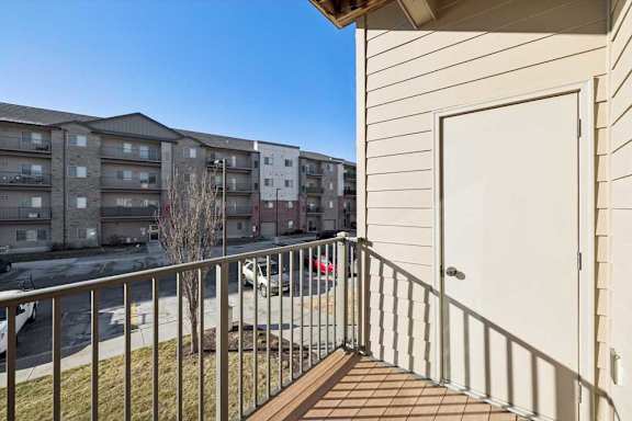 A balcony with a white door and a metal railing overlooks a parking lot and apartment buildings.