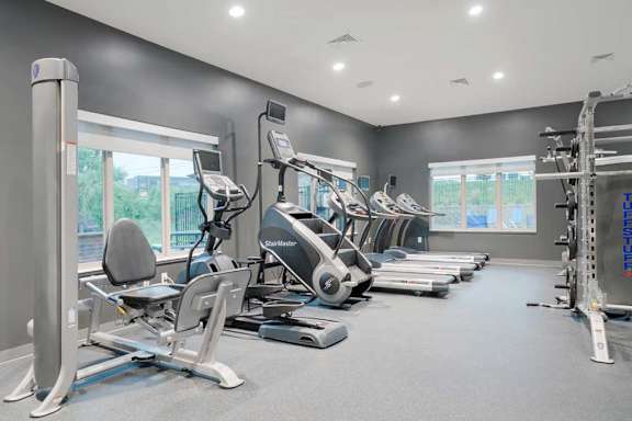 Bright fitness center with cardio machines, large windows, and gray finishes overlooking outdoor community views.