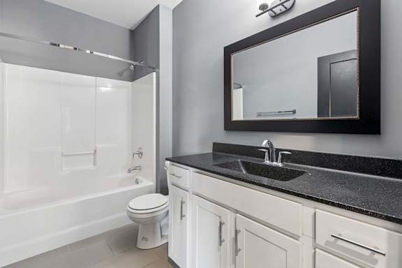 Black-framed mirror and speckled dark countertop contrast with a white vanity and tub-shower combo in this apartment bathroom.