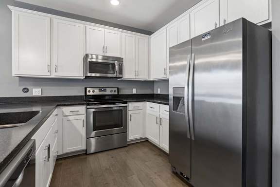 Stainless steel refrigerator, oven, and microwave stand out against crisp white cabinets and dark counters in this apartment kitchen.