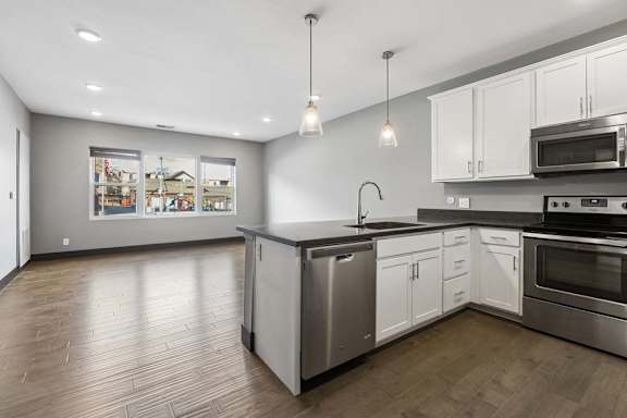 Open-concept apartment kitchen flows into a sunlit living area, with white cabinetry, dark counters, pendant lights, and stainless steel appliances.