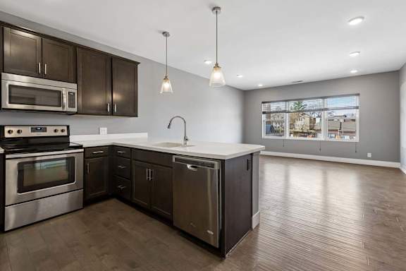 Dark cabinetry and a white peninsula define this open apartment kitchen, flowing into a living area with wide windows and wood-look floors.