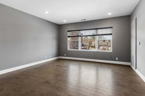 Expansive apartment living room features wood-look flooring, gray walls, and a broad window overlooking the surrounding community.