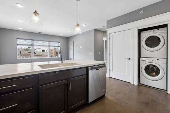 Stacked in-home laundry sits just off the kitchen, beside a dark cabinet island with a deep sink, dishwasher, and pendant lights.