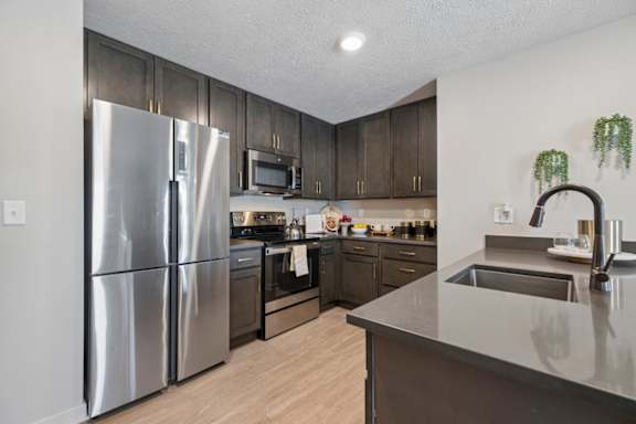 Modern apartment kitchen with dark wood cabinets, stainless steel appliances, light wood flooring, and a large island with sink and countertop seating area.