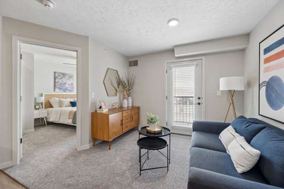 Bright apartment living room with a blue sofa, large window with blinds, neutral carpeting, and an open layout leading to a bedroom and private balcony door.