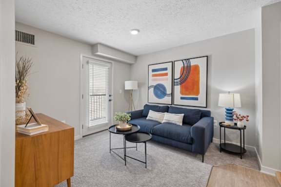 Bright apartment living room with a blue sofa, gray carpet, and a window with blinds providing natural light.
