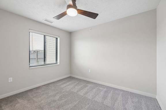 Apartment bedroom with ceiling fan, large window, and neutral carpeting.