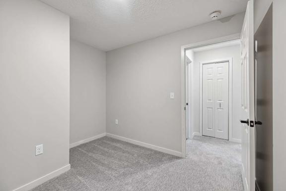 Apartment bedroom with neutral carpeting, light gray walls, and adjacent hallway access.
