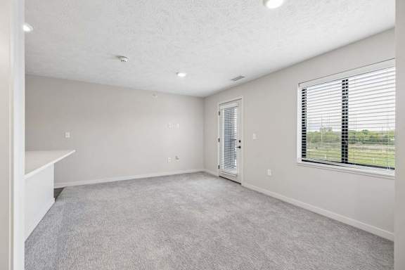 Apartment living area with neutral carpeting, large window, and glass door leading to a private balcony.