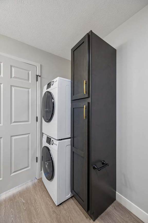 Stacked front-load washer and dryer beside a tall dark storage cabinet in a bright apartment laundry closet with a paneled door and light flooring.