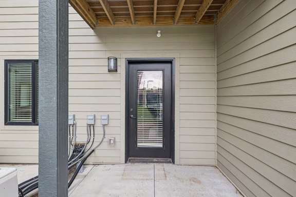 Covered ground-floor apartment patio with a concrete surface, exterior wall lighting, and a glass-paneled door leading into the residence.