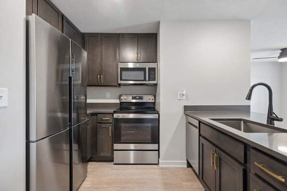 Modern apartment kitchen with dark cabinetry, stainless steel appliances, a deep undermount sink with a pull-down faucet, and a functional open layout.