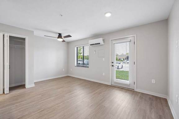 Bright apartment living room with wood-style flooring, recessed lighting, a ceiling fan, a glass patio door, and a wall-mounted mini-split heating and cooling unit.