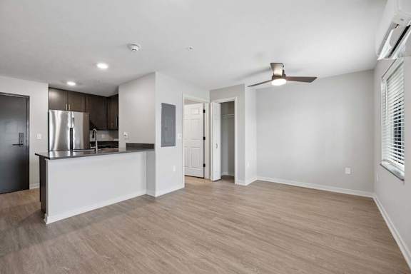 Bright open-concept apartment living room with wood-style flooring, recessed lighting, a ceiling fan, and a kitchen featuring dark cabinetry, stainless steel appliances, and a breakfast bar.