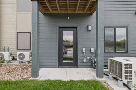 Covered ground-floor apartment patio with a concrete surface, glass-paneled entry door, exterior wall light, and landscaped surroundings.